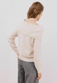 Pull léger beige à épaules dénudées avec finitions côtelées et manches longues, fabriqué à partir d'un tissu doux. Porté avec un jean gris taille haute.