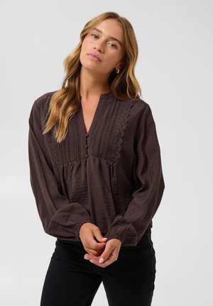 Mulher com cabelo loiro comprido a vestir uma blusa castanha escura com detalhes em renda e botões, em pé com as mãos juntas à frente contra um fundo claro.
