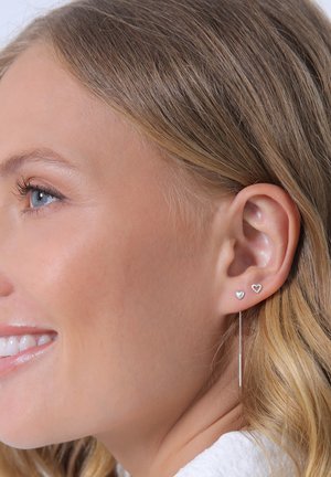 Silberne Ohrringe mit einem Herz-Stecker und einem langen, dünnen Stift mit einem hängenden Akzent, präsentiert vor natürlicher Haartextur und Hautfarbe.