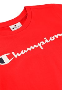 Piros pamut póló nagy fehér "Champion" logóval a mellkason, kerek nyakkivágással és szabványos fazonnal. A címke "S" méretet mutat.