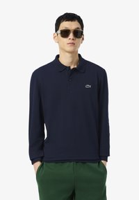 Polo shirt à manches longues de couleur marine avec une texture douce et un col, arborant un petit logo lacoste blanc sur la poitrine, associé à un pantalon vert.