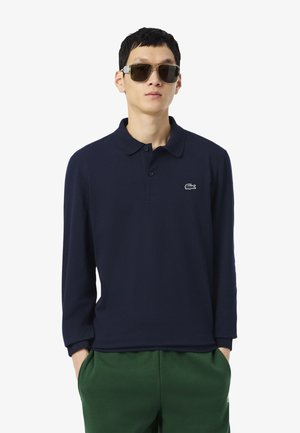 Granatowa koszula polo z długim rękawem o miękkiej teksturze i kołnierzyku, z małym białym logo Lacoste na piersi, zestawiona z zielonymi spodniami.