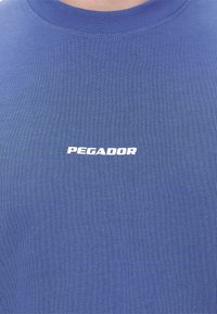 Camiseta azul de cuello redondo con texto blanco "PEGADOR" centrado en el pecho, mostrada desde el cuello hasta la parte superior del torso.