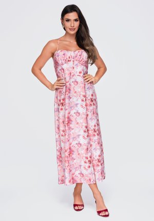Donna che indossa un vestito lungo rosa e rosso a fiori con spalline sottili e tacchi aperti rossi, in piedi con le mani sui fianchi su sfondo bianco.