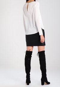 Valkoinen pitkähihainen paita, jossa on takana avainaukko, yhdistettynä muotoon tehtyyn mustaan nahkahameeseen ja reiteen yltävään mustaan mokkanahkasaappaat.