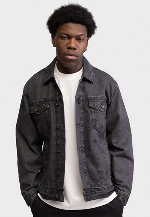 Jeune homme aux cheveux bouclés portant une veste en jean gris foncé par-dessus une chemise blanche, debout devant un fond clair uni.