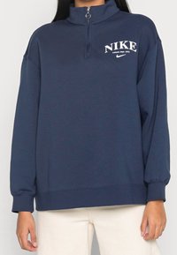 Mörkblå zip-up huvtröja i mjuk bomull, med hög krage, långa ärmar och vitt Nike-logotyp på bröstet.