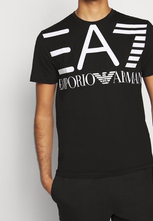 Persona que lleva una camiseta negra con un gran logo blanco "EA7" y "Emporio Armani" impreso en el frente.