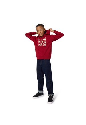 Felpa rossa con cappuccio e motivo geometrico sul davanti, abbinata a jeans blu scuro e sneakers nere. Tessuto liscio e vestibilità comoda.