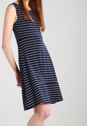 Vrouw die een mouwloze navyblauwe jurk met witte horizontale strepen draagt, staand tegen een effen lichte achtergrond.