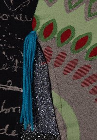 Textil-lapp med svart tyg, vit text, gröna och röda mönster, glänsande silvertextur och en blå kvastdetalj.