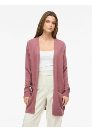 Mujer con un cárdigan abierto color rosa polvoriento sobre una camiseta blanca y pantalones de cintura alta color crema, de pie frente a un fondo liso.