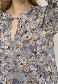 Kwiatowa bluzka z lekkiego, półprzezroczystego materiału. Posiada dekolt w serek, detal z wiązaniem oraz wzór w różnokolorowe kwiaty na szarym tle.