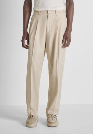 Uomo che indossa pantaloni beige a pieghe, scarpe beige abbinate e una canotta bianca a coste, in piedi contro uno sfondo semplice.