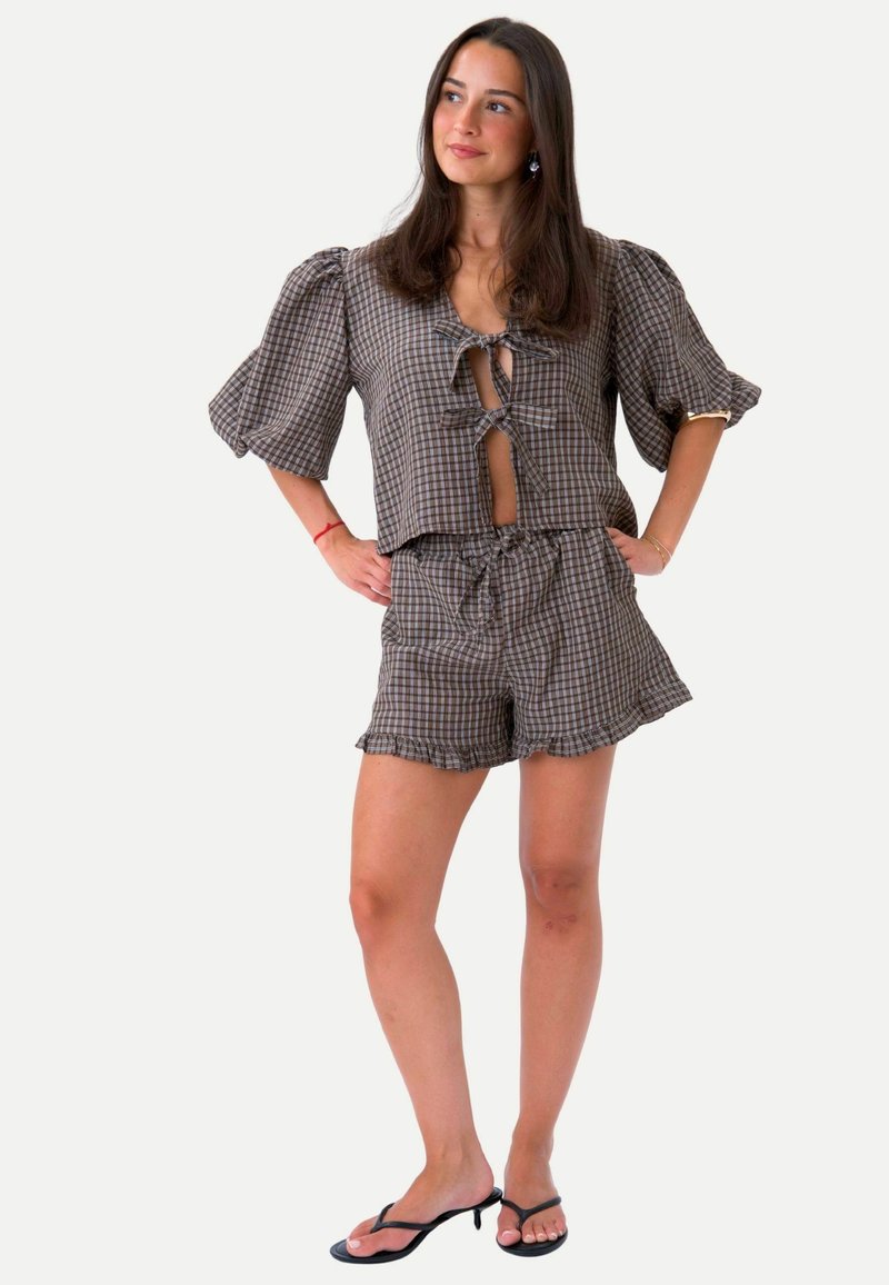 Femme aux longs cheveux portant un haut à manches bouffantes à carreaux marron avec des liens sur le devant, un short assorti à volants et des sandales à talons noires.