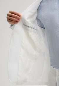 Weißer Blazer mit einer glatten Außenfläche und einem strukturierten Futter mit hexagonalem Muster. Eine versteckte Innentasche mit grauem Akzent ist sichtbar.