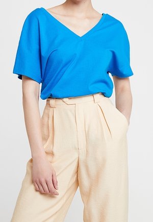 Osoba ubrana w jasnoniebieską koszulkę z krótkim rękawem i dekoltem w serek, wpuszczoną w wysokie, szerokie beżowe spodnie, z jedną ręką w kieszeni.