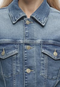 Άτομο φοράει ανοιχτό γαλάζιο τζιν μπουφάν με μεταλλικά κουμπιά και δύο τσεπάκια με κουμπιά στο στήθος· φαίνεται από τους ώμους μέχρι το πάνω μέρος του στήθους.