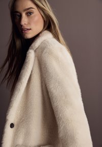 Manteau en fausse fourrure crème avec un col cranté, fermeture par boutons et texture douce. Présente une coupe décontractée. L'arrière-plan est de couleur marron neutre.