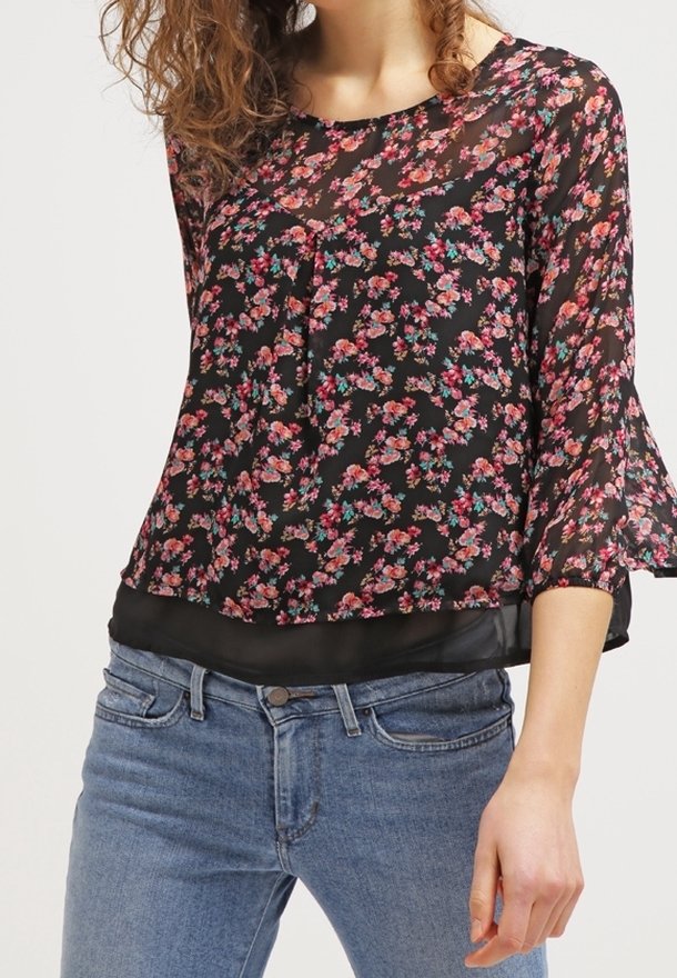 Femme portant un chemisier noir transparent à imprimé floral rose avec des manches cloche, associé à un jean en denim bleu.