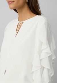 Chemisier blanc avec un col en V et deux fines lanières, doté de manches à volants superposés et d'une texture lisse et légère.