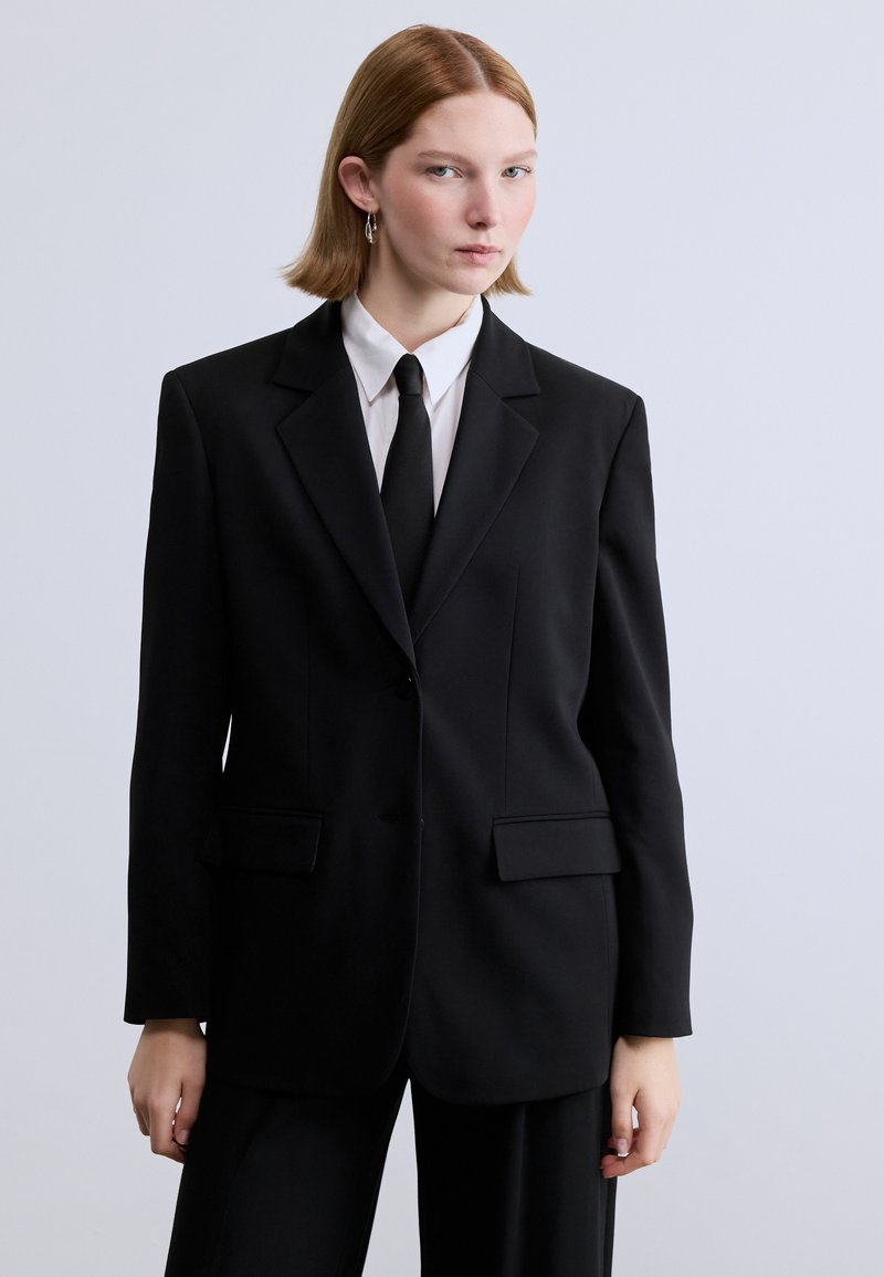 Εξατομικευμένο μαύρο σακάκι με κούμπωμα ενός κουμπιού, δύο μπροστινές τσέπες και πέτο με εγκοπή, φορεμένο πάνω από λευκό πουκάμισο και μαύρη γραβάτα.