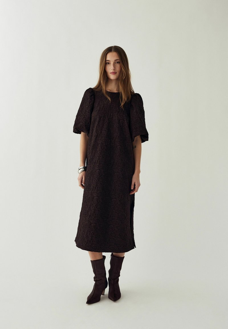 Femme aux cheveux longs portant une robe midi noire texturée avec manches bouffantes et des bottes foncées, debout devant un fond clair uni.