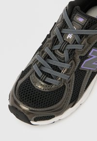 Černá sportovní bota s prodyšným svrškem z meshové látky a lesklými syntetickými doplňky, šedými tkaničkami a bílou mezipodrážkou. Výrazný fialový detail loga.