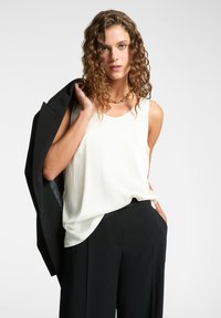 Top bianco senza maniche in una vestibilità rilassata, abbinato a pantaloni neri. La modella tiene un blazer nero sulla spalla, con una finitura leggermente lucida.