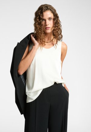 Top bianco senza maniche in una vestibilità rilassata, abbinato a pantaloni neri. La modella tiene un blazer nero sulla spalla, con una finitura leggermente lucida.