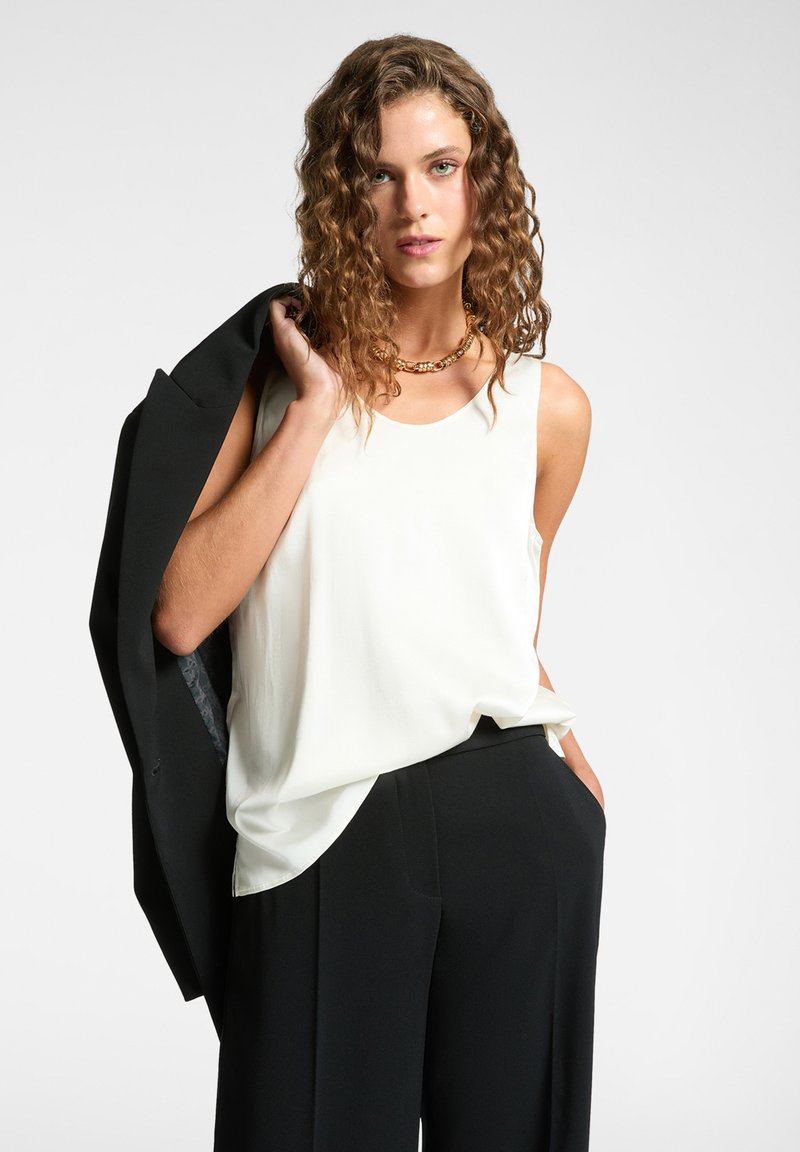 Top bianco senza maniche in una vestibilità rilassata, abbinato a pantaloni neri. La modella tiene un blazer nero sulla spalla, con una finitura leggermente lucida.