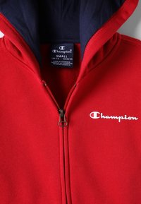 Rode zip-up hoodie met een marineblauwe binnenkant, met een zilveren rits en een witte "Champion" logo op de borst. Zachte stoftextuur.