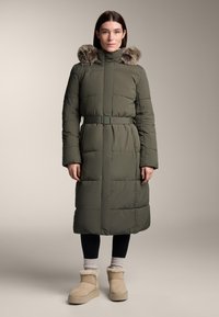 Padded dlhý kabát v olivovej zelenej farbe s vysokým golierom a falošnou kožušinou. Má opasok v páse a prešitú štruktúru. Nosí sa s béžovými kožušinovými čižmami.