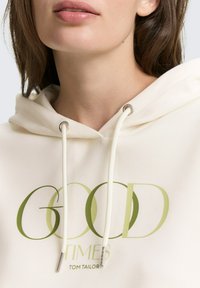 Cremefarbener Hoodie mit einem Frontgrafik in Grün, auf der "GOOD TIMES" steht, ausgestattet mit metallbeschichteten Kordeln und einer weichen Textur.