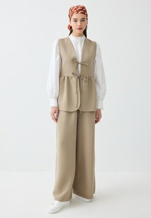 Donna che indossa un gilet beige e pantaloni a gamba larga abbinati a una camicetta bianca, foulard con fantasia, orecchini a cerchio dorati e sneakers bianche.