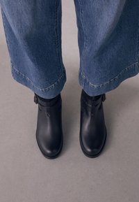 Black leather ankle boots with a smooth finish, featuring buckled straps and rounded toes, paired with loose dark denim jeans.