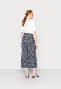 Top branco de manga curta combinado com calças culotte azul marinho com padrão floral. Vista parcial de sandálias rasas e uma tatuagem no tornozelo.