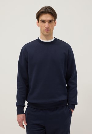 Man draagt een marineblauwe trui en bijpassende broek, staand met één hand in de zak tegen een effen lichte achtergrond.