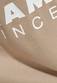 Beige stof med en glat overflade, der har hvid, fed skrift. Designet inkluderer ordet "FAMOUS" og et delvist udsnit af "INCE".