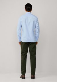 Camisa de manga larga azul claro con puños abotonados, combinada con pantalones verde oscuro y zapatos marrones. Tejido suave, corte ajustado, vista posterior.