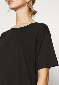 Femme portant un t-shirt noir uni à manches courtes avec col rond, boucle d'oreille en or, et cheveux attachés, sur un fond blanc.