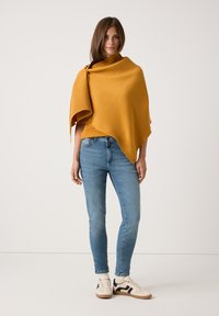 Eine Frau steht da und trägt einen senfgelben Schal, hellblaue Skinny Jeans, weiße Sneakers und cremfarbene Socken vor einem schlichten hellen Hintergrund.