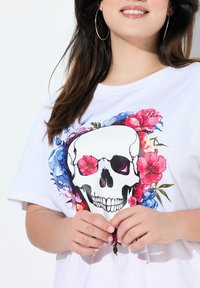 Camiseta blanca con un gráfico central de calavera rodeado de diseños florales coloridos en rosa, azul y verde. Manga corta y corte relajado.