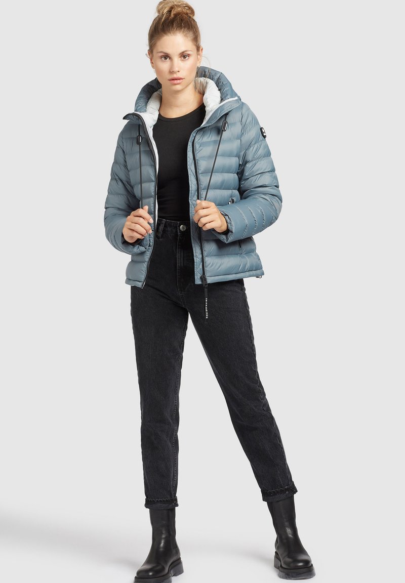 Hellblaue Daunenjacke mit weißem Fleecefutter, mit Reißverschluss und Kordelzugkapuze, kombiniert mit schwarzen Jeans und Stiefeln.