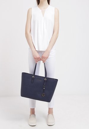 Borsa a tracolla blu marina con design strutturato, due manici robusti, dettagli in hardware dorato e un charm pendente. Indossata con una cima bianca e pantaloni bianchi.