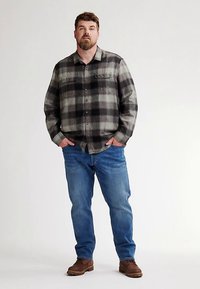 Camicia di flanella a quadri neri e grigi con due tasche sul petto, abbinata a jeans azzurri e scarpe marroni. Tessuto uniforme e vestibilità casual.