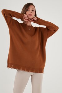 Sieviete valkā vaļīgu brūnu džemperi ar nodilušām malām un bēšas krāsas bikses, pozējot ar rokām pie sejas pret vienkāršu fonu.