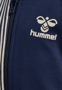 Navy zip-up jacket made of textured fabric, featuring white vertical stripes and a prominent white hummel logo on the chest.
