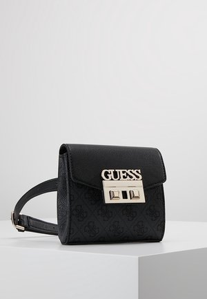 Schwarze Guess-Gürteltasche mit strukturierter Klappe und silberner Schnalle mit Markenlogo, die auf einem weißen Sockel vor einfarbigem Hintergrund liegt.