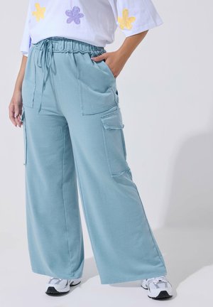 Persona che indossa pantaloni cargo larghi azzurri con tasche a toppa e scarpe da ginnastica bianche, con una mano in tasca, in piedi davanti a uno sfondo neutro.